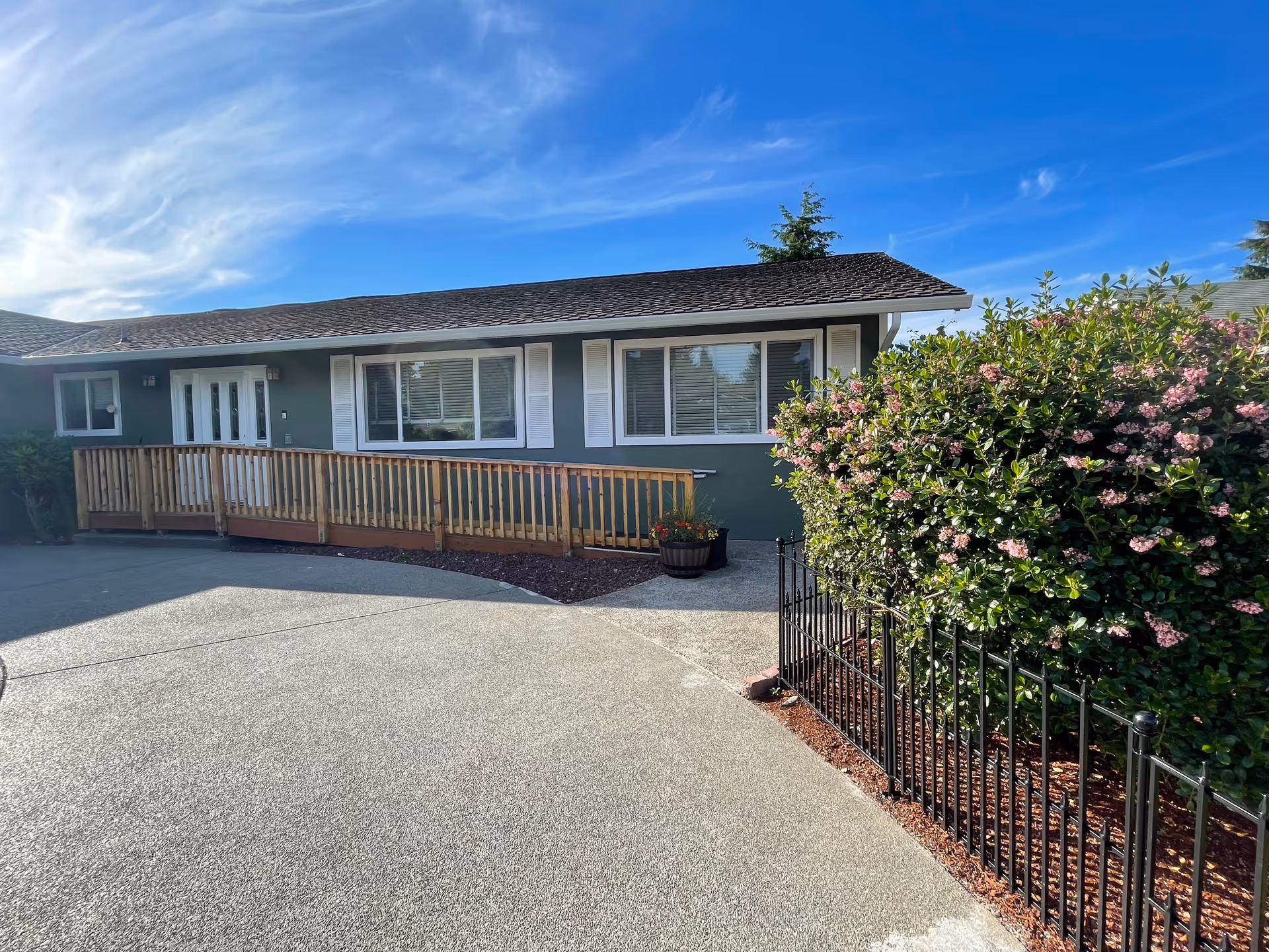 Exterior view of a single-story building with a wooden ramp leading to a white double door entrance. The building has green walls with white window shutters and large windows. There is a black metal fence with flowering bushes on the right side and a clear blue sky with some clouds above.