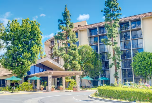 Exterior view of a multi-story senior living facility with balconies, surrounded by tall trees and greenery under a clear blue sky. The entrance features a covered drop-off area with outdoor seating and green umbrellas.