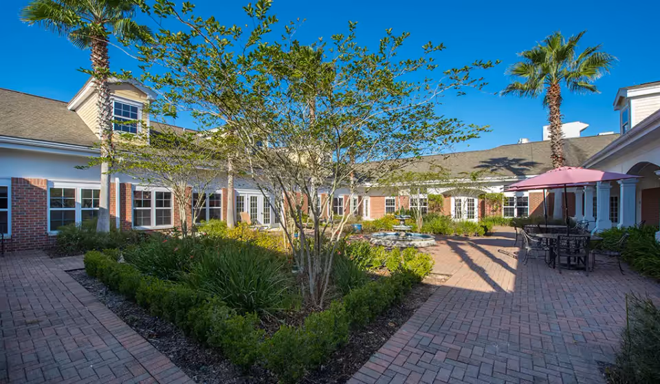 Courtyard with brick pathways, a planted garden and fountain surrounded by a one-story red-brick building, palm trees, and outdoor seating with a pink umbrella.