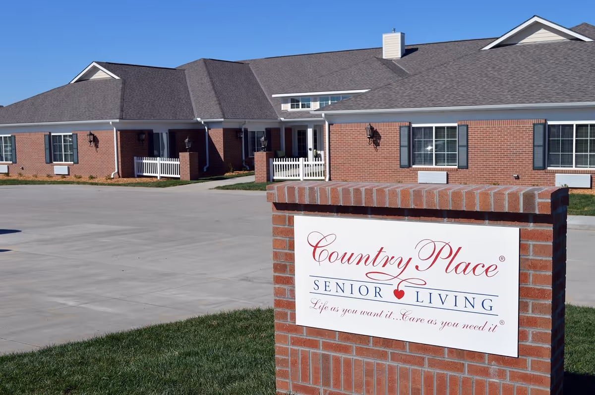 Exterior view of a single-story brick senior living facility with a paved driveway and a large sign in the foreground that reads 'Country Place Senior Living Life as you want it... Care as you need it'. The building has multiple windows with shutters and a clear blue sky above.