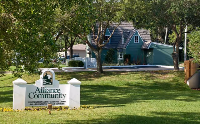 A green residential-style building surrounded by trees and grass with a white sign in the foreground that reads 'Alliance Community Independent Living - Assisted Living Skilled Nursing - Rehab Services'.