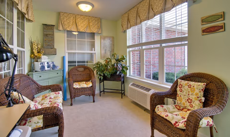 Sunlit sitting room with wicker chairs and floral cushions, a small cabinet, plants, and large windows.