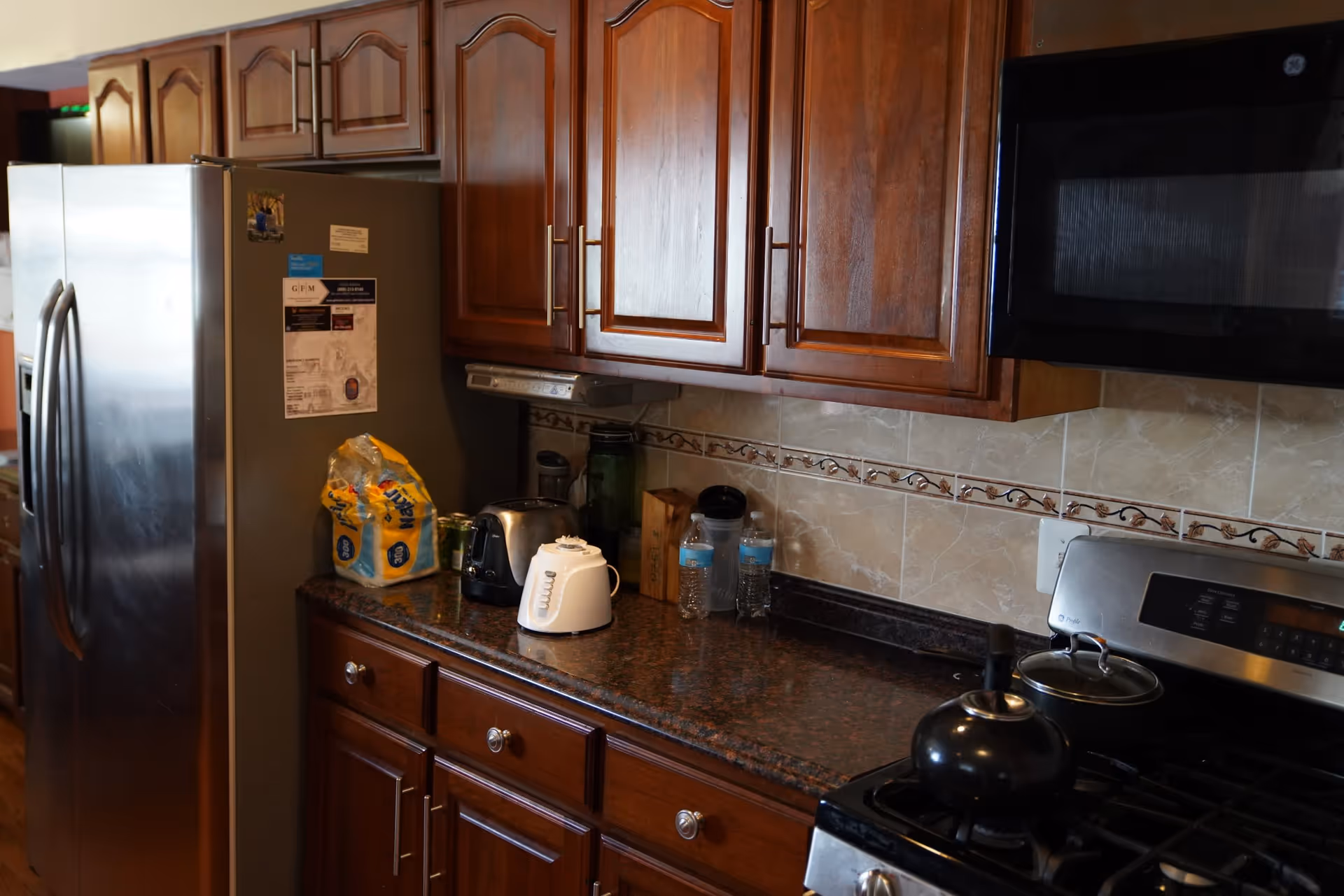 Kitchen area with wooden cabinets, a stainless steel refrigerator, a microwave, a stove with a kettle and pot, and various items on the countertop including a toaster, a white electric kettle, water bottles, and a bag of bread.