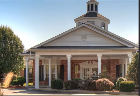 Front exterior view of a senior living facility building with white columns, brick walls, and a cupola on the roof. There are bushes and trees around the entrance under a clear blue sky.