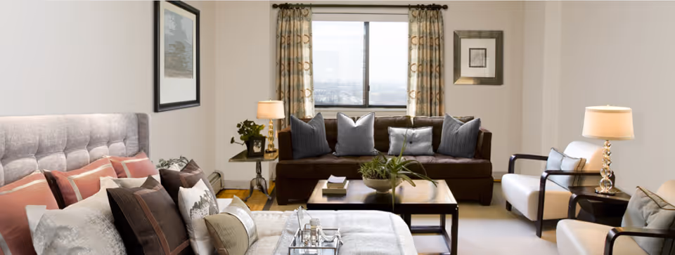 A cozy living room area with a large window in the background. The room features a brown sofa with multiple gray and blue cushions, two white armchairs with cushions, a coffee table with a plant and books, and a bed with various pillows in the foreground. There are two table lamps on side tables and framed artwork on the walls.