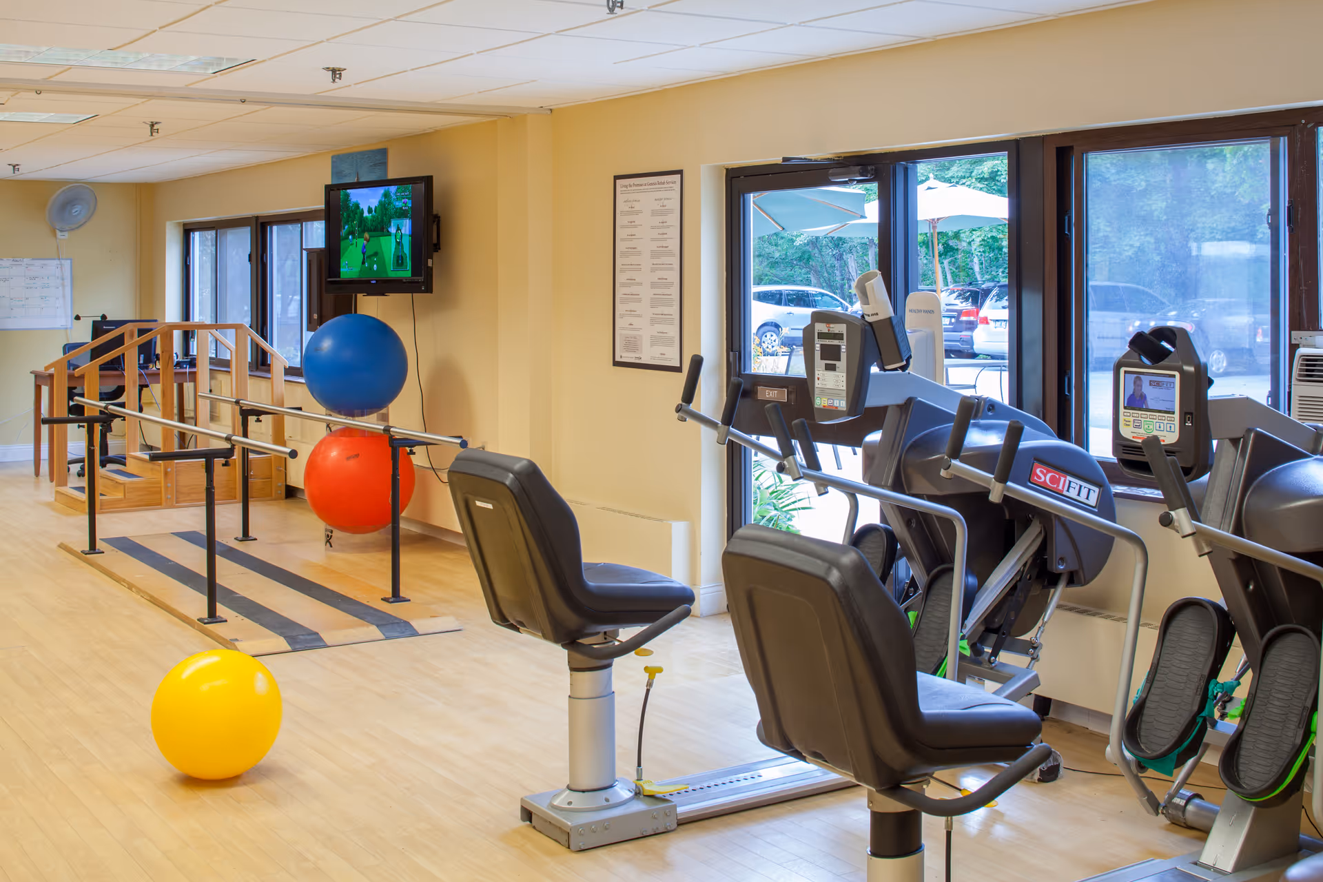 Interior view of a senior living facility exercise room with parallel bars for walking support, colorful exercise balls, and seated exercise machines near large windows showing parked cars outside. A television is mounted on the wall displaying a golf game.