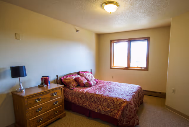 Small furnished bedroom with a double bed in patterned red bedding, a wooden nightstand with a lamp, and a window letting in daylight.