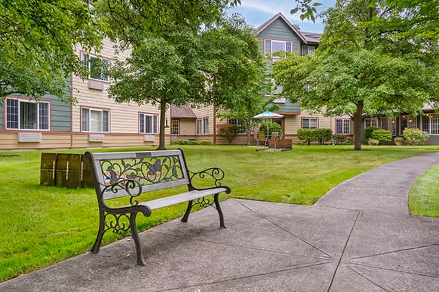 Outdoor courtyard area of a senior living facility with a decorative metal bench on a concrete pathway, green grass, trees, and a multi-story building with beige and green siding in the background.