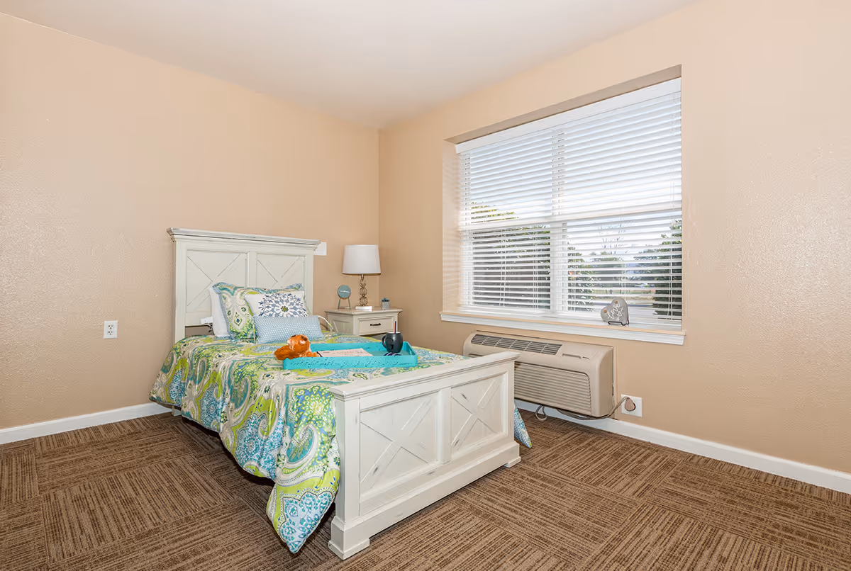 A small bedroom with a single white wooden bed featuring a colorful green and blue patterned bedspread. There are several pillows on the bed, a small stuffed animal, and a blue tray with a cup on it. Next to the bed is a white nightstand with a lamp, a clock, and a small plant. A large window with white blinds is on the right wall, and below it is an air conditioning unit. The walls are beige, and the floor is carpeted in a brown pattern.
