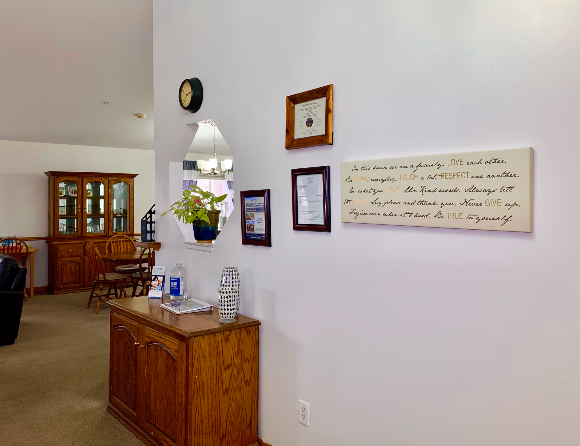 Interior view of a senior living facility showing a wooden cabinet with a plant, hand sanitizer, and a decorative vase on top. On the wall above the cabinet are framed certificates and a decorative sign with inspirational text. In the background, there is a dining area with wooden chairs and a china cabinet.