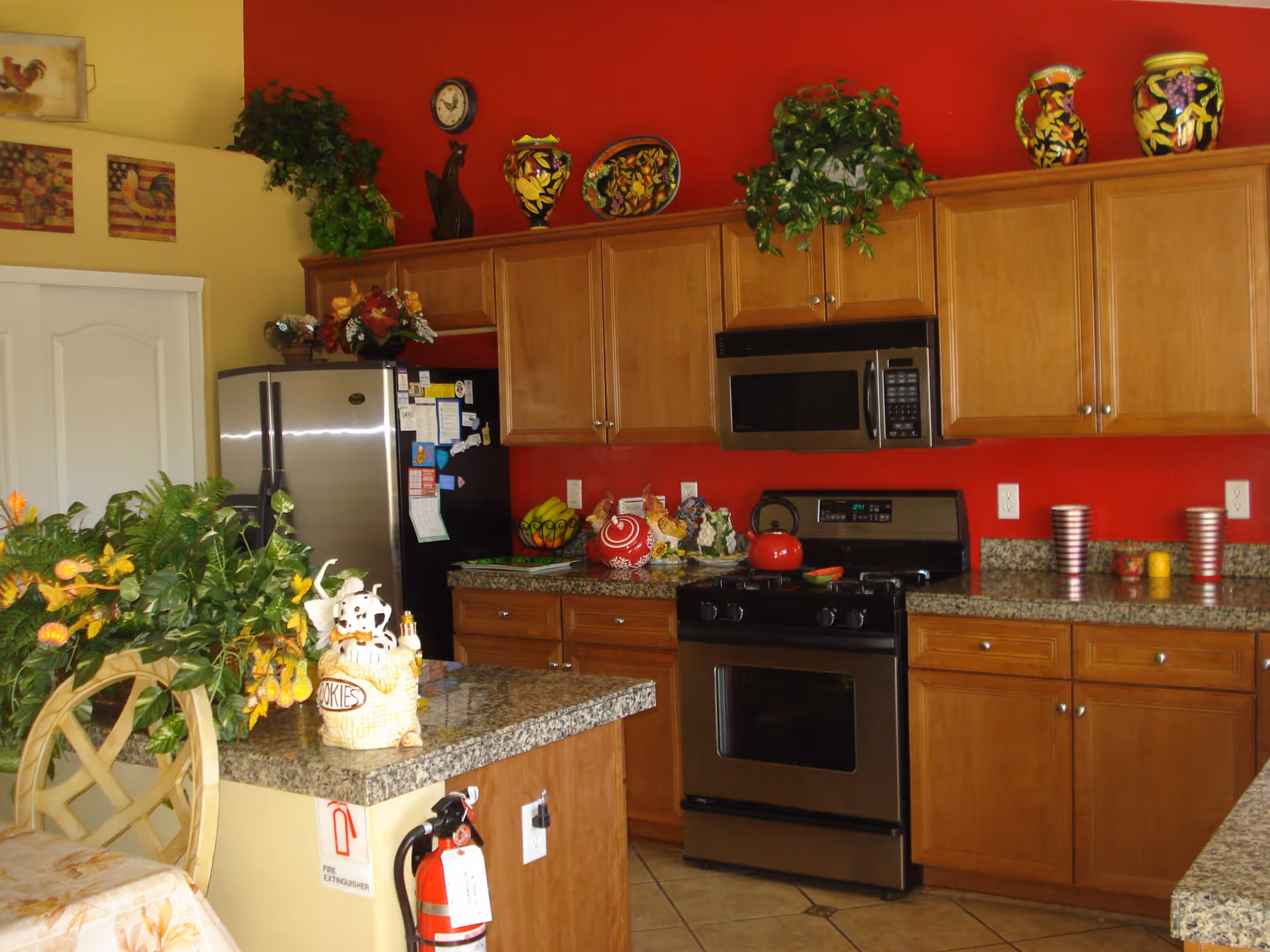 A kitchen with wooden cabinets, granite countertops, and stainless steel appliances including a refrigerator, microwave, and stove. The walls are painted red and yellow, decorated with plants, colorful ceramic vases, and rooster-themed artwork. A fire extinguisher is mounted on the side of the kitchen island, which also holds a decorative cookie jar and a floral arrangement.