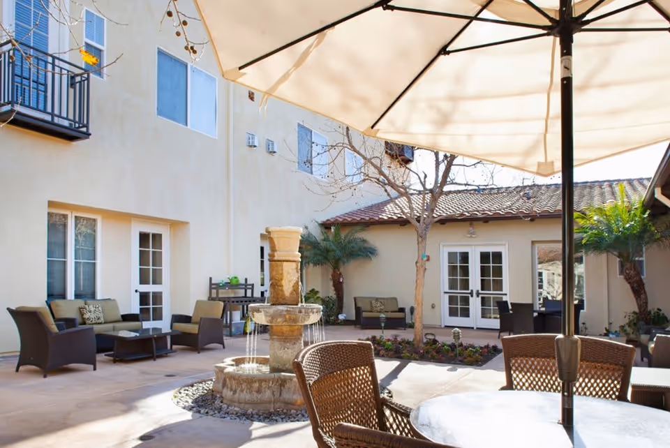 Outdoor courtyard area with a stone water fountain in the center, surrounded by patio furniture including wicker chairs and cushioned sofas. There are potted plants, small trees, and a large umbrella providing shade over a table. The courtyard is enclosed by a beige building with multiple windows and doors.