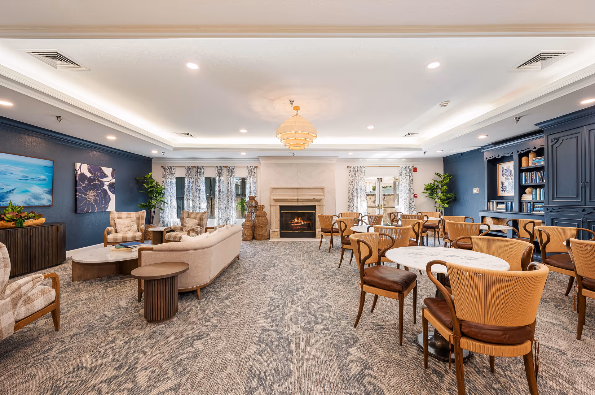 Bright communal lounge and dining area with round tables and chairs and a seating area with sofas facing a fireplace.