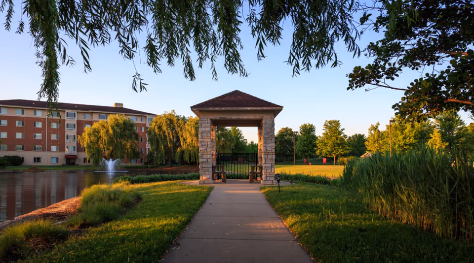 A peaceful outdoor scene at Sedgebrook featuring a paved walkway leading to a small stone pavilion with a peaked roof. The pavilion is situated near a pond with a water fountain, surrounded by lush green grass, trees, and shrubs. A multi-story residential building is visible in the background under a clear sky.