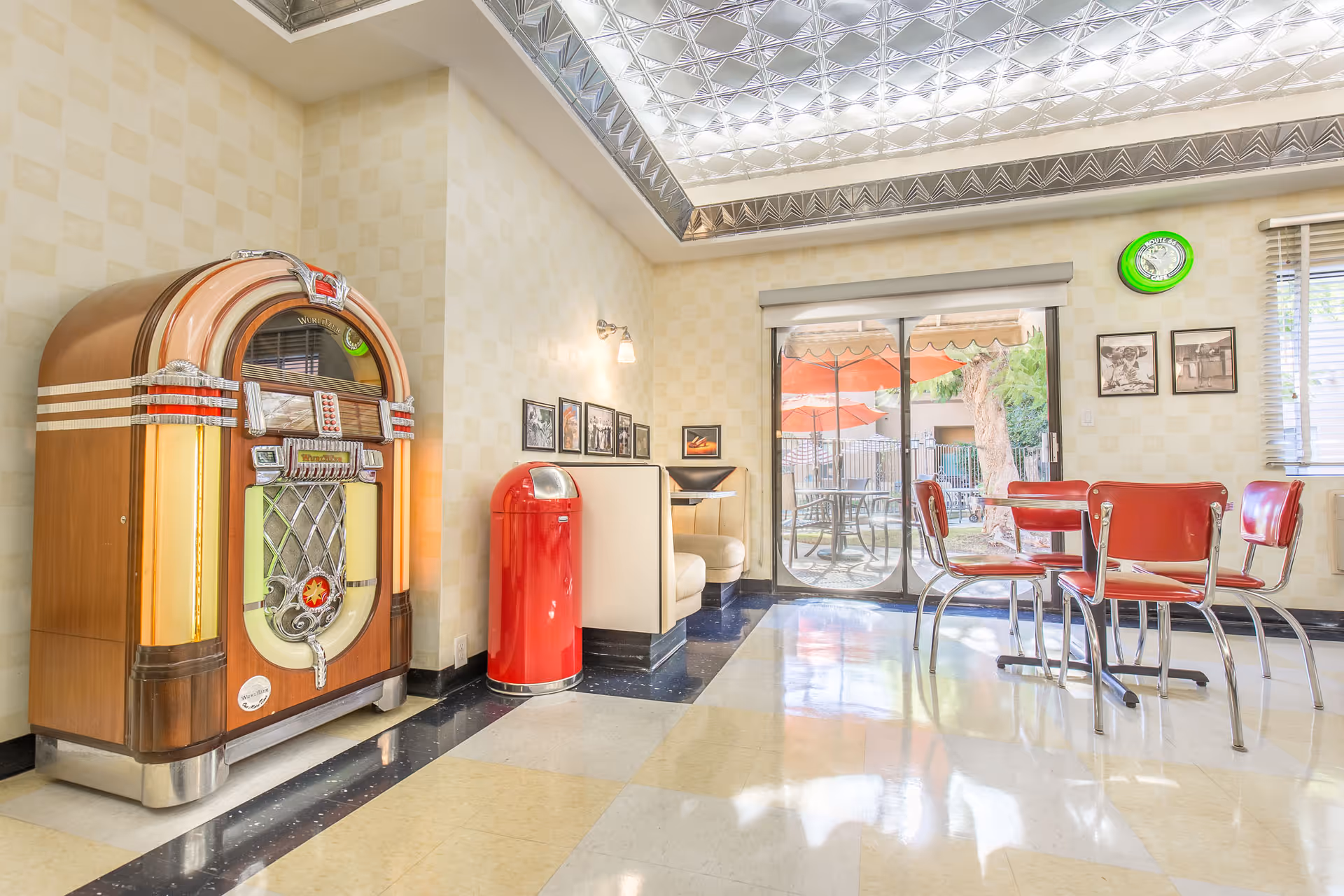A retro-style interior room featuring a vintage jukebox, a red trash bin, a small booth seating area, and a dining table with four red chairs. The room has checkered wallpaper, a glass block ceiling, and a sliding glass door leading to an outdoor patio with tables and umbrellas.