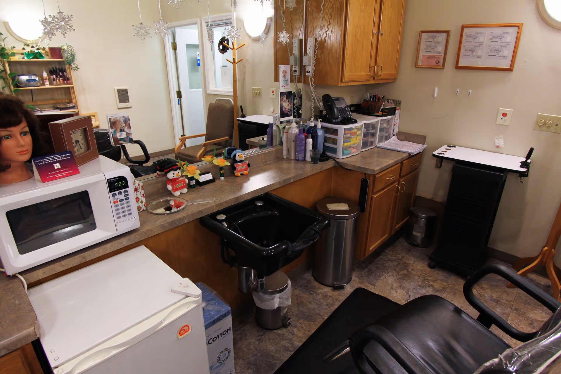 Interior view of a salon area in a senior living facility with a black salon chair, a black hair washing sink, a microwave, a small refrigerator, various hair care products on the counter, a mannequin head with a wig, and a mirror reflecting part of the room. The room has wooden cabinets, a coat rack, and some framed documents on the wall.
