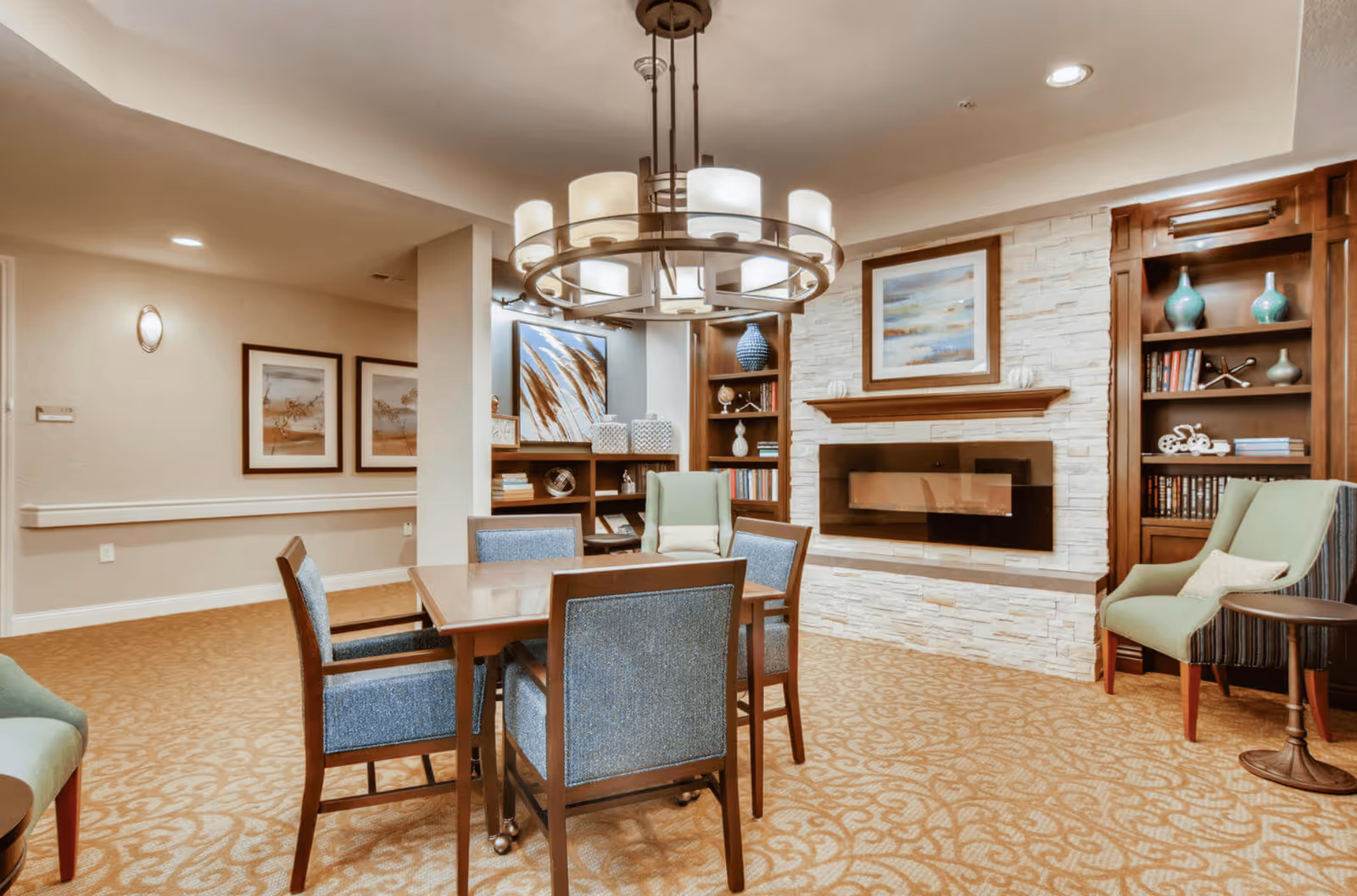 A cozy interior common area with a square wooden table surrounded by four blue upholstered chairs. The room features a modern circular chandelier with multiple lights hanging from the ceiling. In the background, there is a white stone fireplace with a framed abstract painting above it, flanked by wooden bookshelves filled with books and decorative items. Two green armchairs with pillows are positioned near the fireplace, and the carpet has a beige pattern. The walls are decorated with framed artwork and soft lighting.