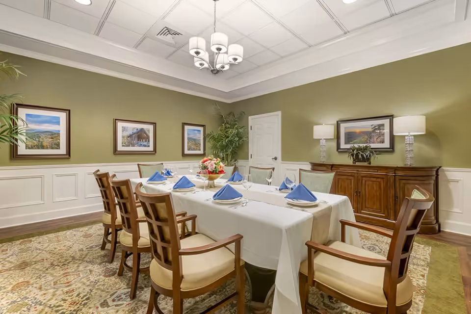 Formal dining room with a long table set with a white tablecloth and blue napkins, wooden chairs, framed artwork, and green walls.