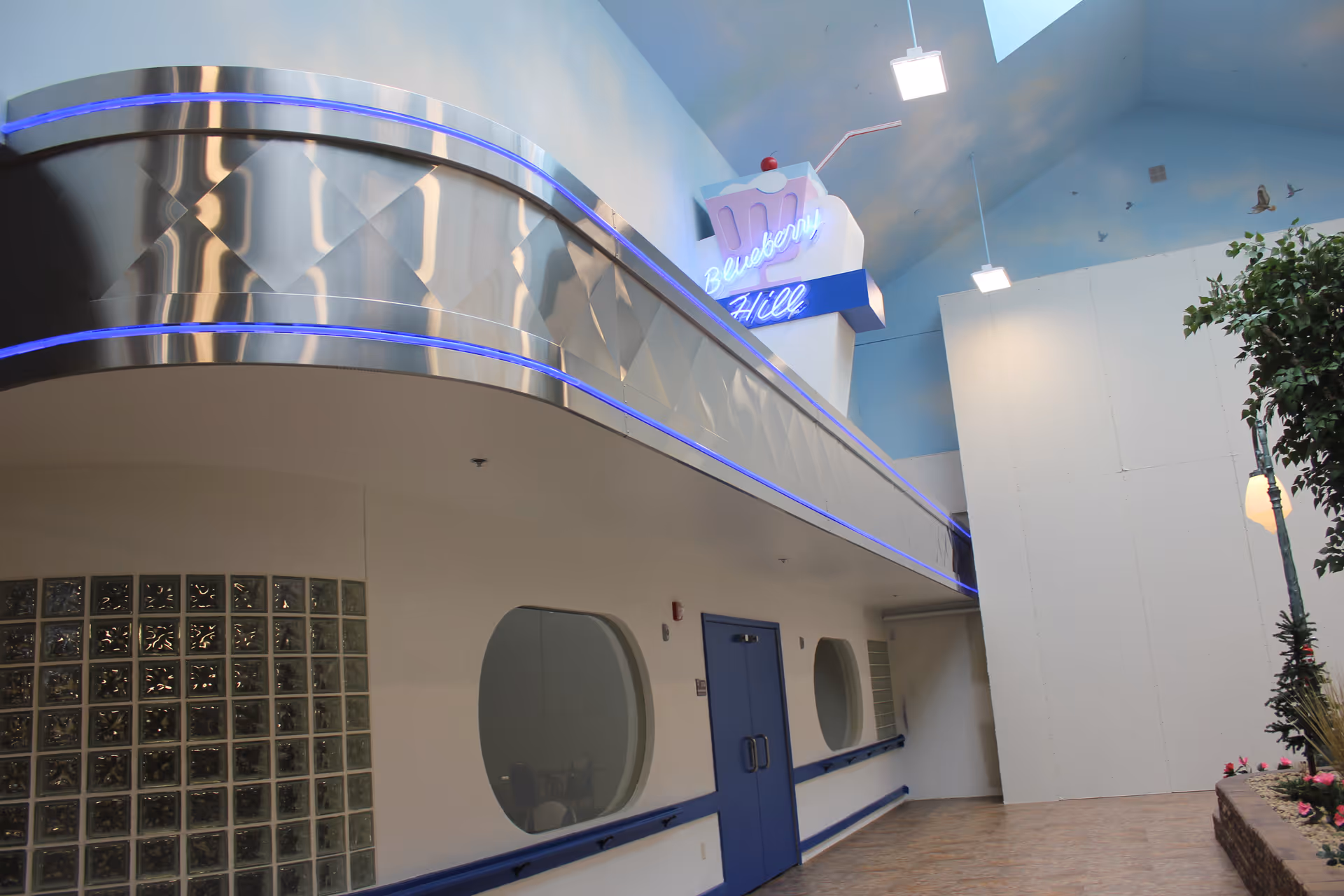 Interior hallway of a facility with a retro diner-style sign reading 'Blueberry Hill' mounted above a metallic balcony with blue neon lights. The hallway has blue double doors, round windows, glass block wall, and a potted tree with flowers on the right side. The ceiling is high with skylights and hanging lights.