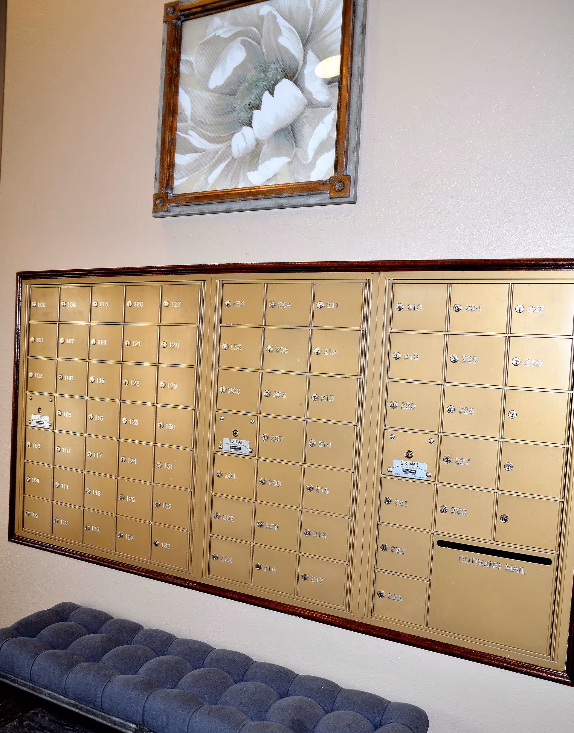 A wall-mounted set of gold-colored mailboxes with numbered compartments, three U.S. Mail slots, and an outgoing mail slot. Above the mailboxes is a framed painting of a large white flower. Below the mailboxes is a tufted blue bench.