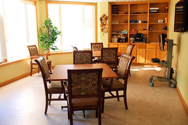 A well-lit room with two wooden tables surrounded by cushioned chairs with patterned upholstery. There is a large window with blinds letting in natural light, a green potted plant in the corner, wooden built-in shelves and cabinets along the back wall, and a mobile TV stand with a flat screen on the right side.