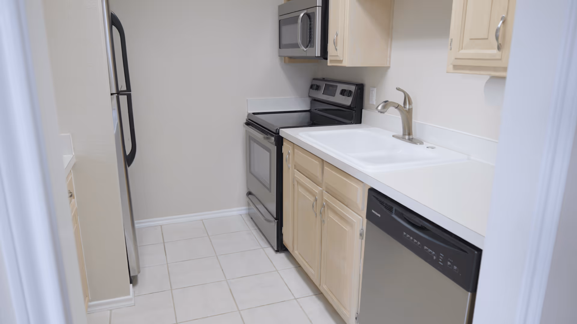 A small kitchen with light wood cabinets, a white countertop, a white sink with a silver faucet, a stainless steel dishwasher, a black and stainless steel oven with a stove, a microwave above the oven, and a stainless steel refrigerator. The floor is tiled with light-colored tiles.