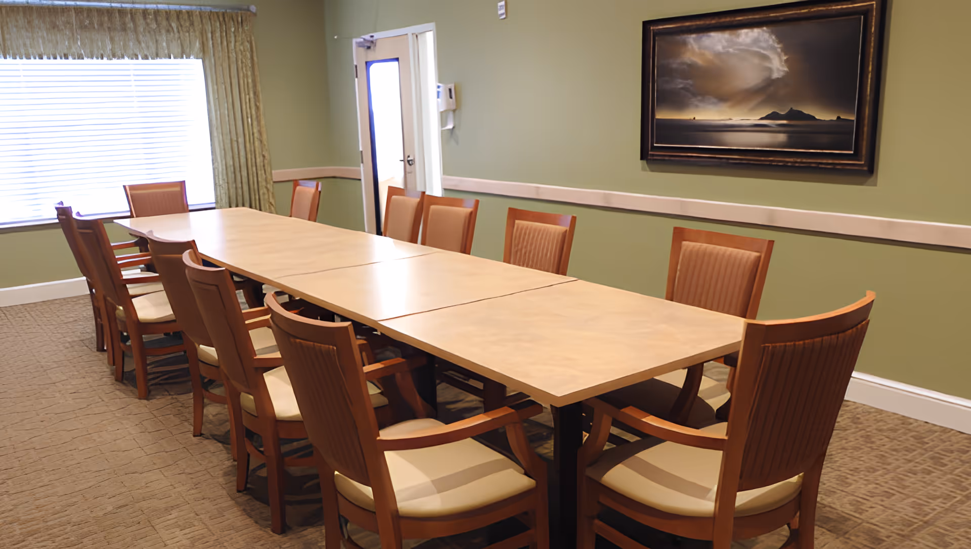 A dining/meeting room with a long rectangular table surrounded by wooden chairs, green walls, a window with blinds, and framed artwork.