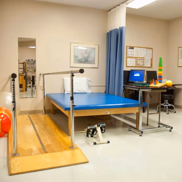 A physical therapy room with a blue padded therapy table, parallel bars on a wooden ramp for walking exercises, a small pedal exerciser on the floor, and a desk with a computer and colorful therapy toys. The room has beige walls, a framed picture, a mirror, and a blue privacy curtain.