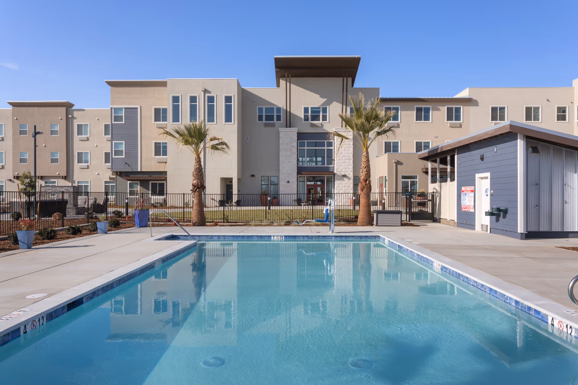 Outdoor swimming pool with clear blue water in front of a multi-story residential building with beige and gray exterior walls. Two palm trees are planted near the pool area, and there is a small building to the right side of the pool. The sky is clear and blue.