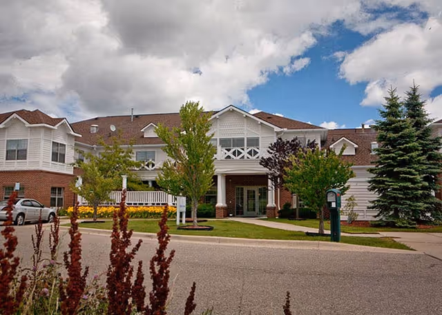 Exterior view of The Cortland Howell senior living facility showing a two-story building with white siding and brick accents, surrounded by trees and landscaping under a partly cloudy sky.