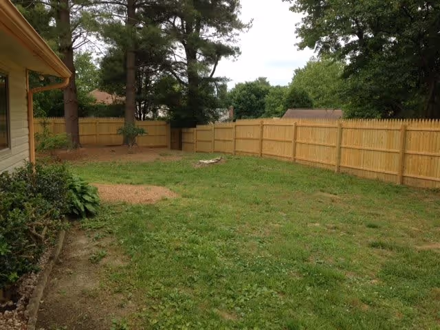 Fenced grassy backyard with trees and the side of a house visible on the left.