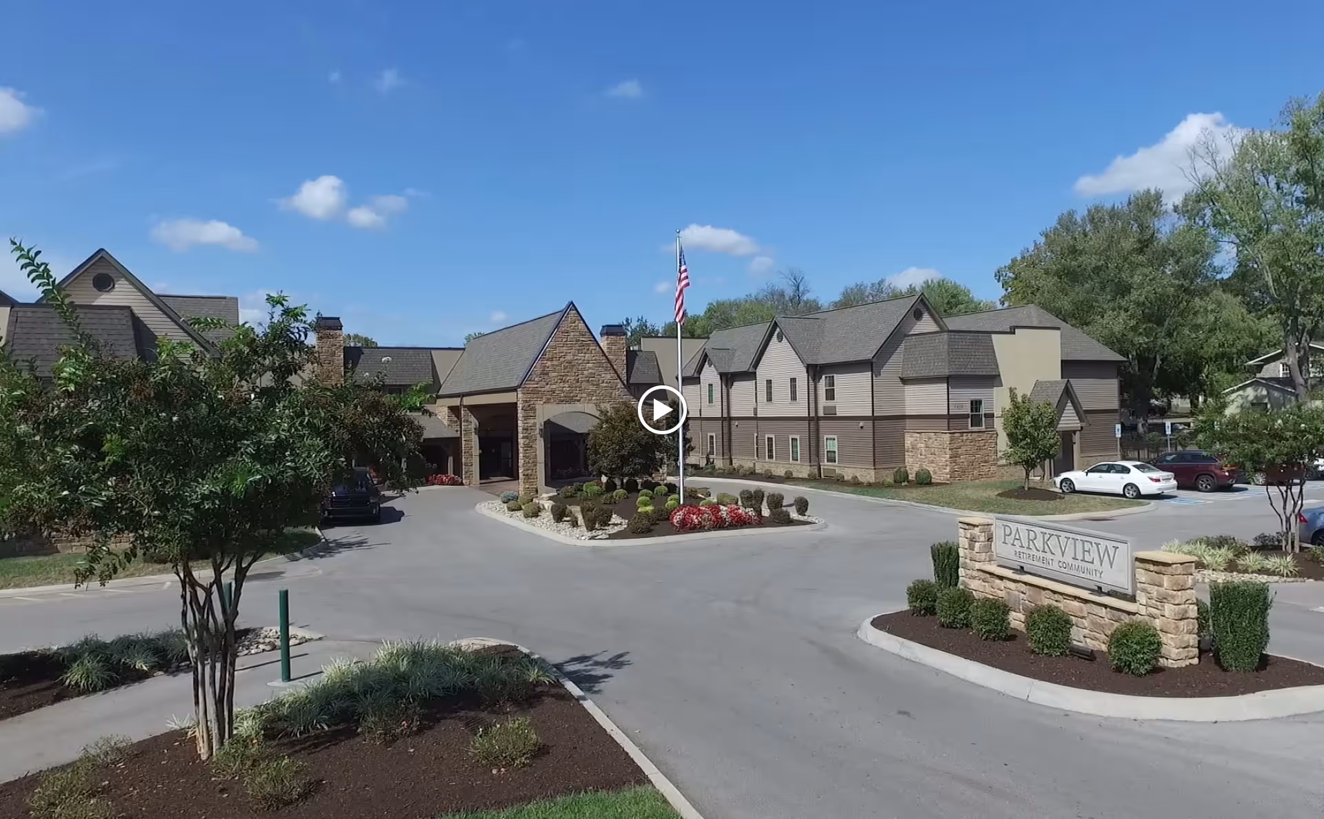 Exterior view of Parkview Senior Living - West Knoxville facility showing a two-story building with a stone and siding facade, a driveway with landscaped islands, an American flag on a flagpole, and a sign reading 'Parkview Retirement Community' at the entrance.