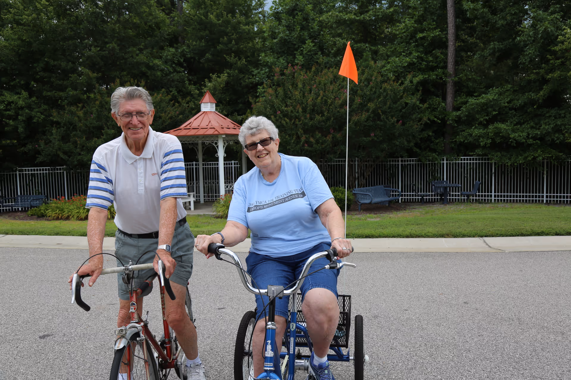 An elderly man and woman smiling while riding bicycles on a paved path in an outdoor area with green trees, a gazebo, and benches in the background. The woman is on a tricycle with an orange safety flag.
