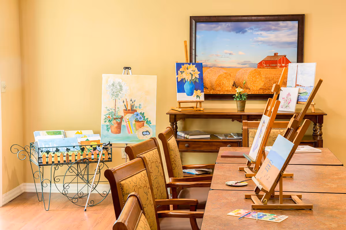 Bright interior activity room with tables and chairs set up with easels and paintings, plus artwork displayed on a side table and wall.