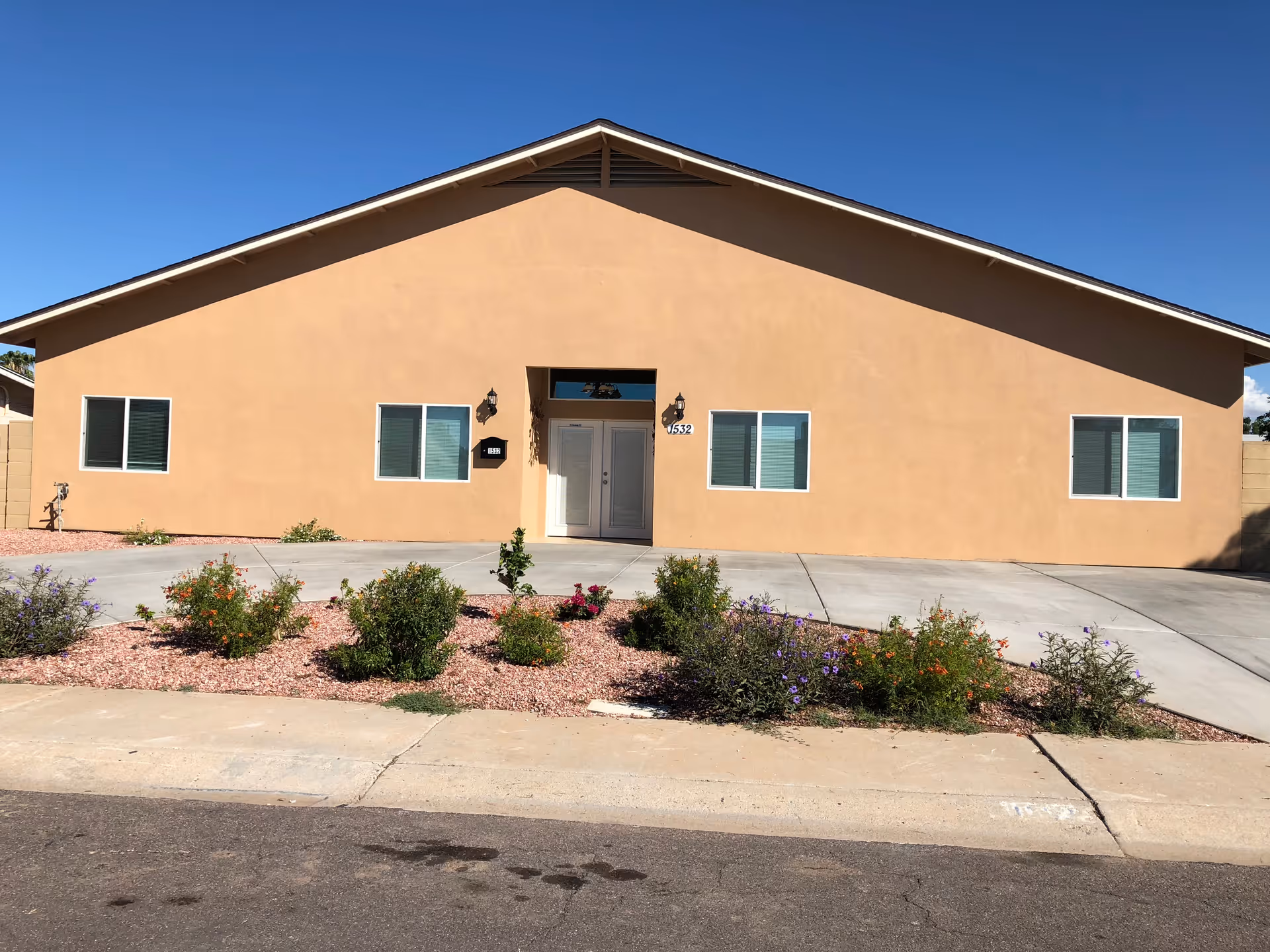 Front exterior view of a single-story building with a beige stucco finish, four windows, a central double-door entrance, and a small garden with various plants and flowers in front. The sky is clear and blue.