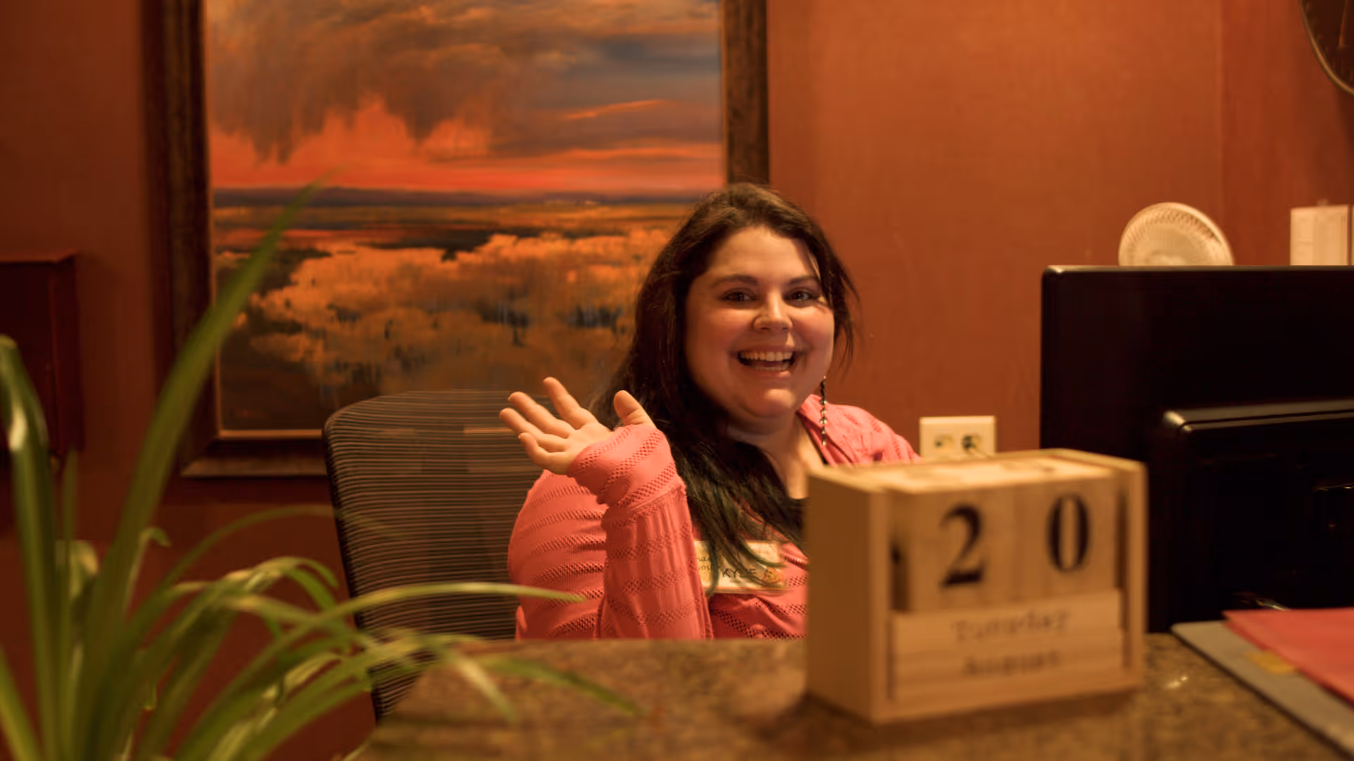 A smiling woman with long dark hair wearing a pink sweater waves while sitting behind a reception desk. On the desk is a wooden block calendar showing the date Tuesday, August 20. There is a plant in the foreground and a landscape painting with warm colors on the wall behind her.
