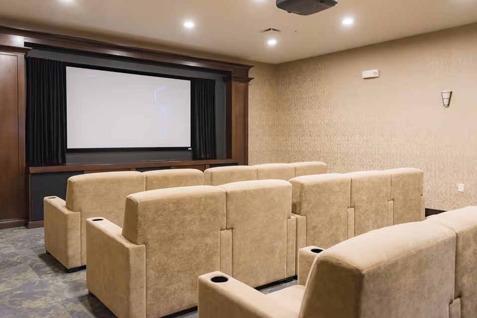 A small theater room with beige upholstered reclining chairs arranged in rows facing a large blank projection screen framed by dark wood paneling. The walls are covered with patterned wallpaper and the ceiling has recessed lighting and a projector mounted.