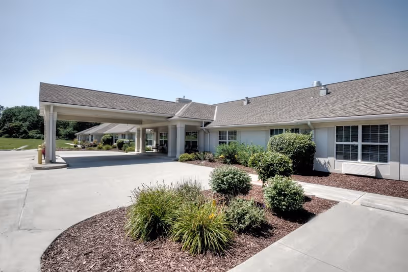 Exterior view of a single-story senior living facility building with a covered entrance driveway, landscaped bushes and plants, and clear blue sky.