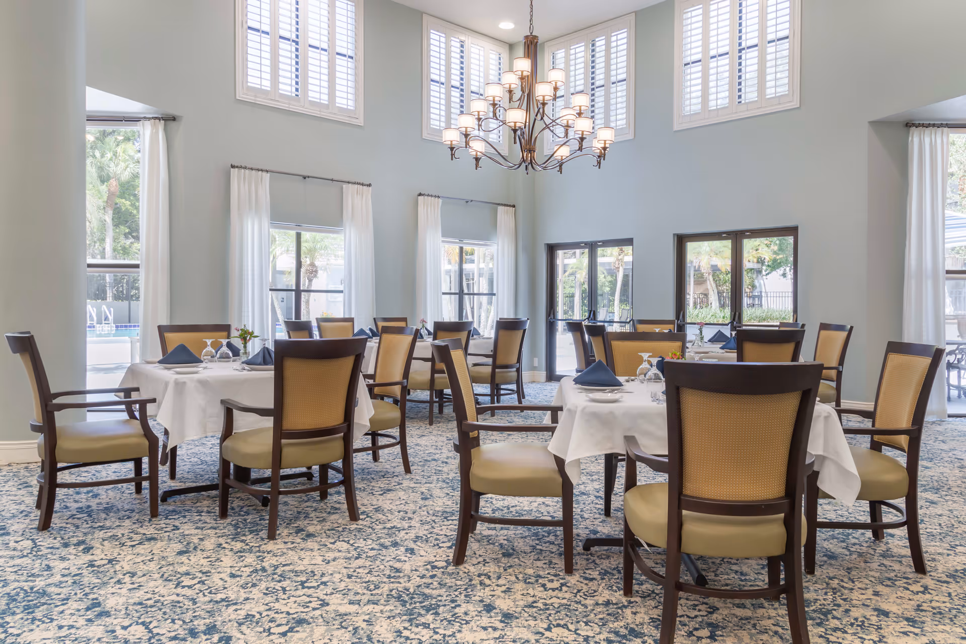 A bright dining room with multiple round tables covered with white tablecloths, each set with plates, glasses, and folded napkins. The room features high ceilings with large windows and white curtains, allowing natural light to fill the space. A large chandelier hangs from the ceiling, and outside the windows, palm trees and a pool area are visible.