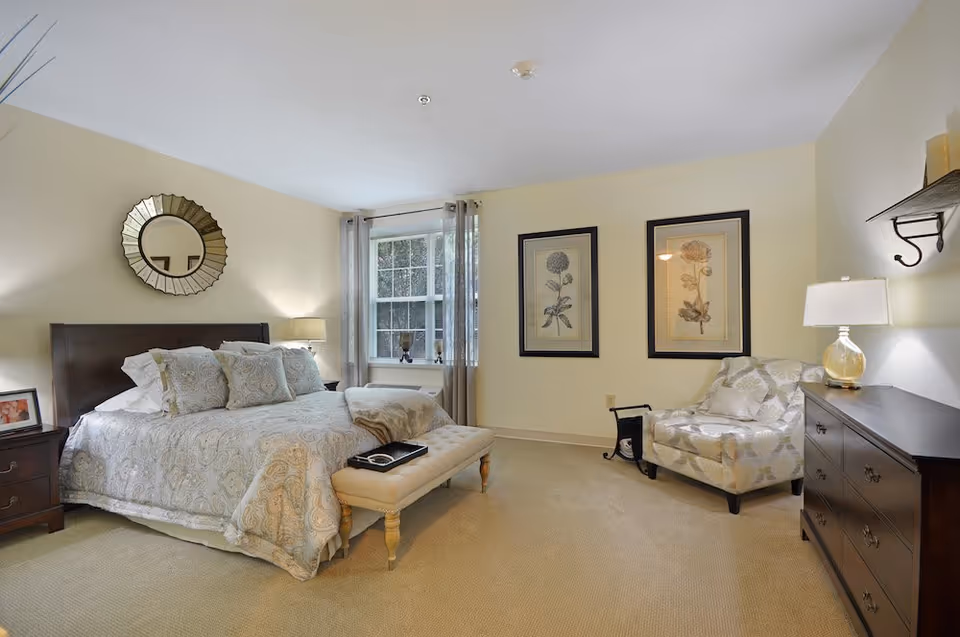 A cozy bedroom with a large bed adorned with patterned bedding and multiple pillows. There is a cushioned bench at the foot of the bed, a nightstand with a lamp and framed photo, and a decorative round mirror on the wall above the bed. A window with sheer curtains lets in natural light. On the opposite wall, two framed botanical prints hang above a patterned armchair next to a dark wooden dresser with a lamp on top.