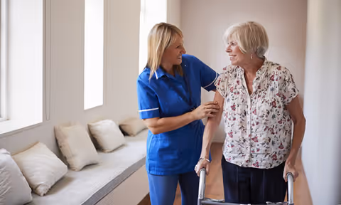 A female caregiver in a blue uniform smiling and assisting an elderly woman using a walker in a bright hallway with a cushioned bench and pillows along the wall.