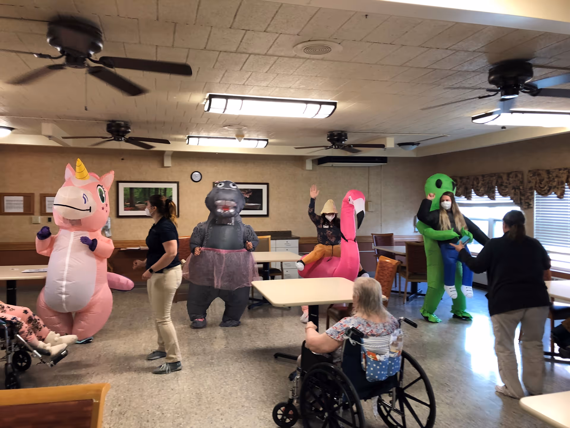 A group of people in a senior living facility common area, with some individuals dressed in inflatable animal costumes including a pink unicorn, a gray dinosaur, a pink flamingo, and a green alien. Two staff members are interacting with residents, one of whom is seated in a wheelchair. The room has ceiling fans, tables, chairs, and framed pictures on the walls.