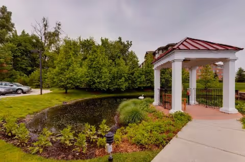 Outdoor area at Fox Run Senior Living Community featuring a small pond surrounded by greenery, a paved walkway, a white gazebo with a red roof, and parked cars in the background under a cloudy sky.