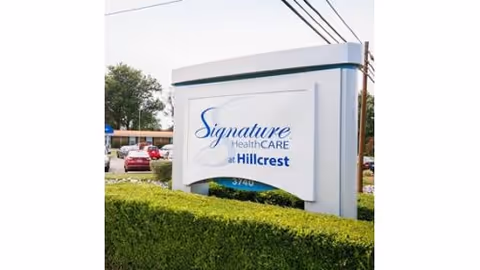 Outdoor sign for Signature HealthCARE at Hillcrest surrounded by green hedges with a parking lot and trees in the background.