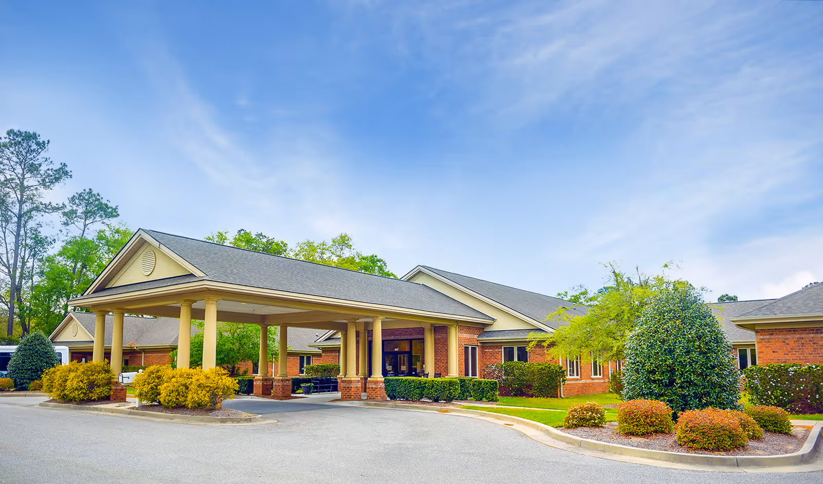Exterior view of Anderson Oaks Assisted Living facility showing a single-story brick building with a covered entrance supported by columns, surrounded by well-maintained landscaping including bushes and trees under a partly cloudy sky.