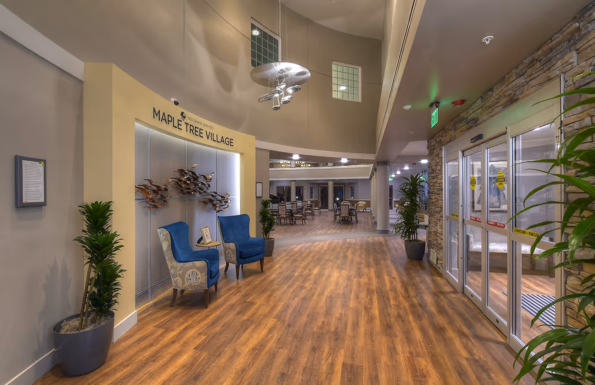 Interior view of the entrance area of ACC Maple Tree Village senior living community featuring a wooden floor, two blue armchairs with a small table between them, decorative wall art, potted plants, and automatic glass doors on the right. In the background, there is a dining area with tables and chairs.
