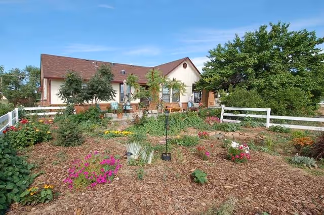 A single-story building with a brown roof and cream-colored walls is visible behind a garden filled with various plants and flowers. The garden is bordered by a white wooden fence, and there are several trees and shrubs around the area under a clear blue sky.