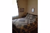 Small bedroom with a double bed covered in a patterned quilt, a nightstand with lamp, and framed pictures on the walls near a curtained window.