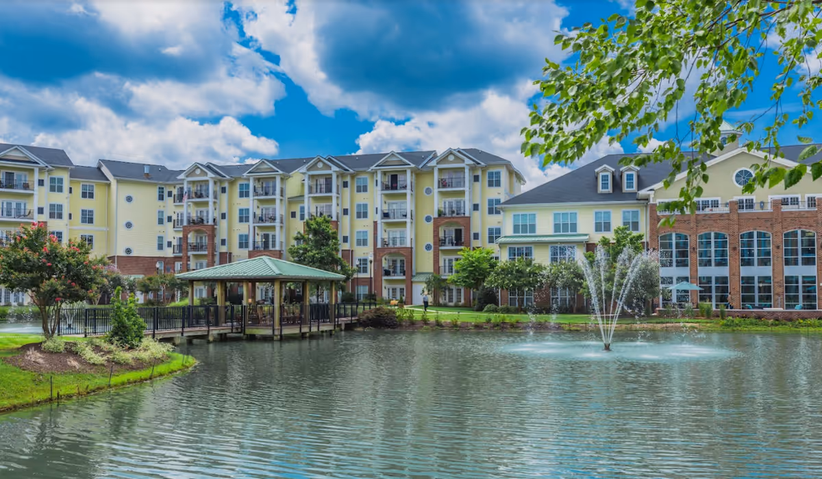 A large senior living facility building with multiple floors and balconies, situated behind a pond with a water fountain. There is a covered wooden gazebo extending over the pond, surrounded by landscaped greenery and trees under a partly cloudy blue sky.