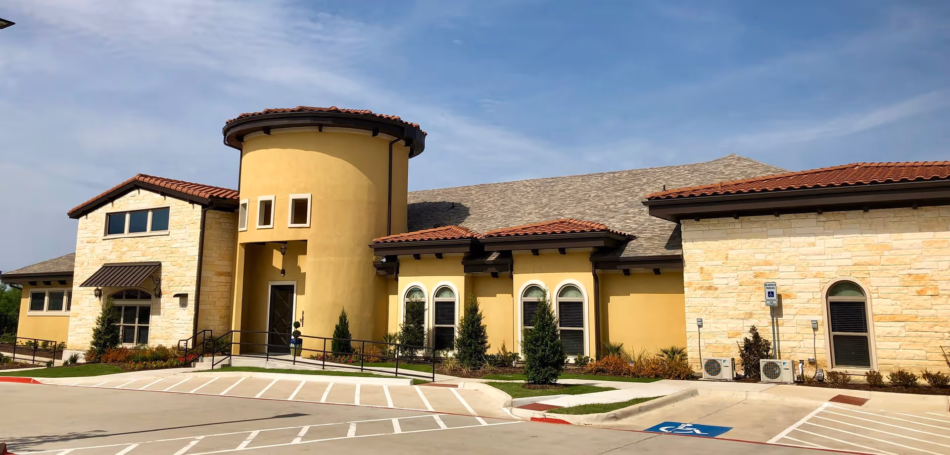 Exterior view of Windhaven Senior Living facility showing a building with a combination of yellow stucco and light stone walls, red tile roof, arched windows, and a round tower-like entrance. The foreground includes a parking lot with marked handicap parking spaces and landscaped greenery around the building.